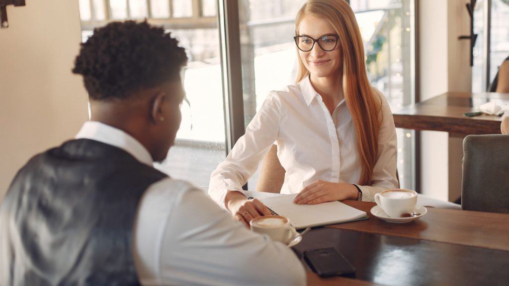 Giving negative feedback at work: A black man wearing business clothes sits across a desk from a white woman in business clothes. She is smiling