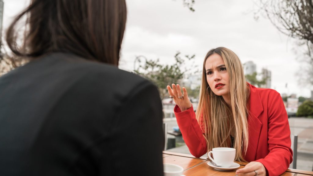 A woman wearing office clothes sits at a picnic table with a coffee. She's angry and gesturing at another coworker sitting across from her. She doesn't know how to deal with defensive coworkers.